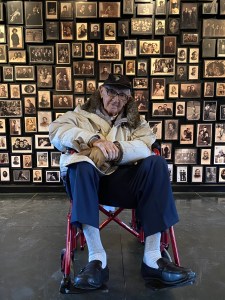 On his visit to the Auschwitz-Birkenau State Museum in 2020, David Wisnia sits before a display of photographs of people imprisoned there during the Holocaust. The display covers a wall of what was once the "sauna" facility of the death camp, where prisoners were given clothing. This was David's workplace during his imprisonment for two and a half years. Photo courtesy Retro Report.